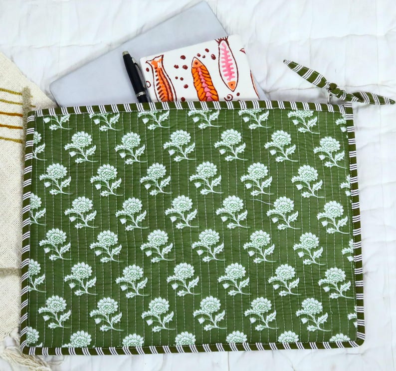 May include: A rectangular quilted laptop sleeve in olive green featuring a repeating white floral pattern. The sleeve has a striped border and a zip closure with a fabric tie. Inside, a silver laptop and a white notebook with fish illustrations.