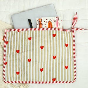 May include: A quilted fabric laptop sleeve featuring a beige and white vertical striped pattern with small red hearts scattered across the surface. The sleeve has a red and white striped border and a matching fabric tie closure on the right side.