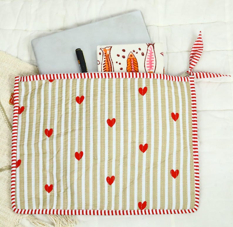 May include: A quilted fabric laptop sleeve featuring a beige and white vertical striped pattern with small red hearts scattered across the surface. The sleeve has a red and white striped border and a matching fabric tie closure on the right side.