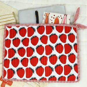 May include: A square quilted fabric pouch featuring a repeating pattern of red strawberries on a white background, finished with red and white striped trim and a matching fabric tie. A black pen and a white card are tucked inside the pouch.