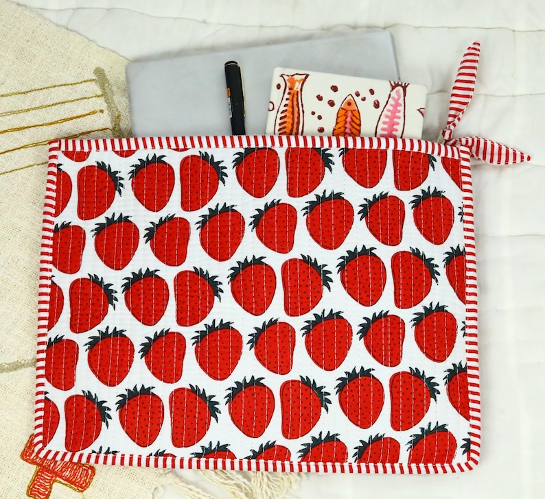 May include: A square quilted fabric pouch featuring a repeating pattern of red strawberries on a white background, finished with red and white striped trim and a matching fabric tie. A black pen and a white card are tucked inside the pouch.
