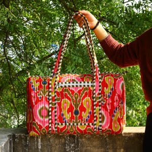 May include: A quilted duffel bag featuring a vibrant red, pink, and yellow ikat pattern. The bag includes a rectangular front pocket with a zipper closure, two long shoulder straps, and a top zipper opening, all finished with a green and white trim.