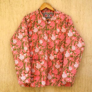 Könnte beinhalten: Eine rosafarbene gesteppte Jacke mit Blumendruck. Die Jacke hat einen Stehkragen und einen Knopfverschluss. Sie hat zwei Taschen auf der Vorderseite.