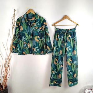 Può includere: Un pigiama verde e blu con stampa giungla con animali e piante. La parte superiore ha una chiusura con bottoni sul davanti e maniche lunghe. I pantaloni hanno una cintura elastica.