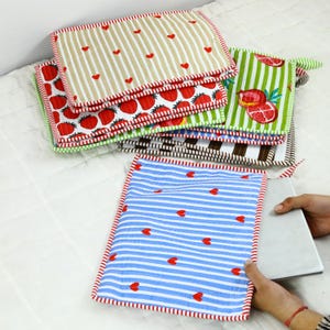 Può includere: Una pila di articoli quadrati in tessuto colorato con vari motivi. I disegni includono cuori rossi su strisce blu, fragole e strisce marroni e bianche. Ogni articolo ha un bordo rosso a contrasto.