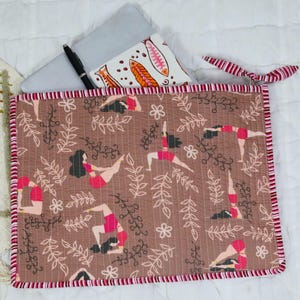May include: A rectangular quilted fabric sleeve featuring a brown background with a repeating pattern of figures in yoga poses, white floral outlines, and leafy branches. The sleeve has a red and white striped fabric trim and a matching fabric tie.