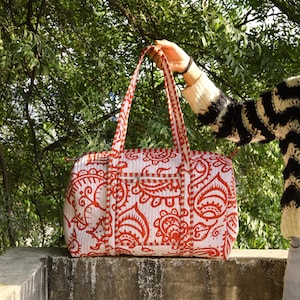 May include: A quilted duffel bag featuring a white base with a vibrant orange paisley pattern. The bag includes two long shoulder straps, a horizontal zippered front pocket, and a top zipper closure, all accented with a matching orange and white striped trim.