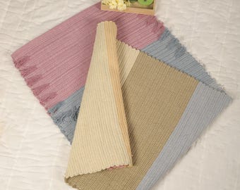 Può includere: Una collezione di tappeti tessuti nei toni del rosa, beige e grigio. Un tappeto è arrotolato, gli altri sono stesi, mostrando un design strutturato con bordi sfrangiati. Un piccolo vassoio di legno con fiori e un vaso verde è sullo sfondo.