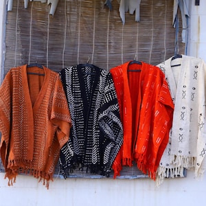 Op de afbeelding: Vier kimono's met patronen in roest, zwart, rood-oranje en crème hangen voor een bamboe jaloezie. Elke kimono heeft een uniek geometrisch ontwerp in contrasterende kleuren en een gefranjerde zoom. De kimono's worden op zwarte hangers getoond.