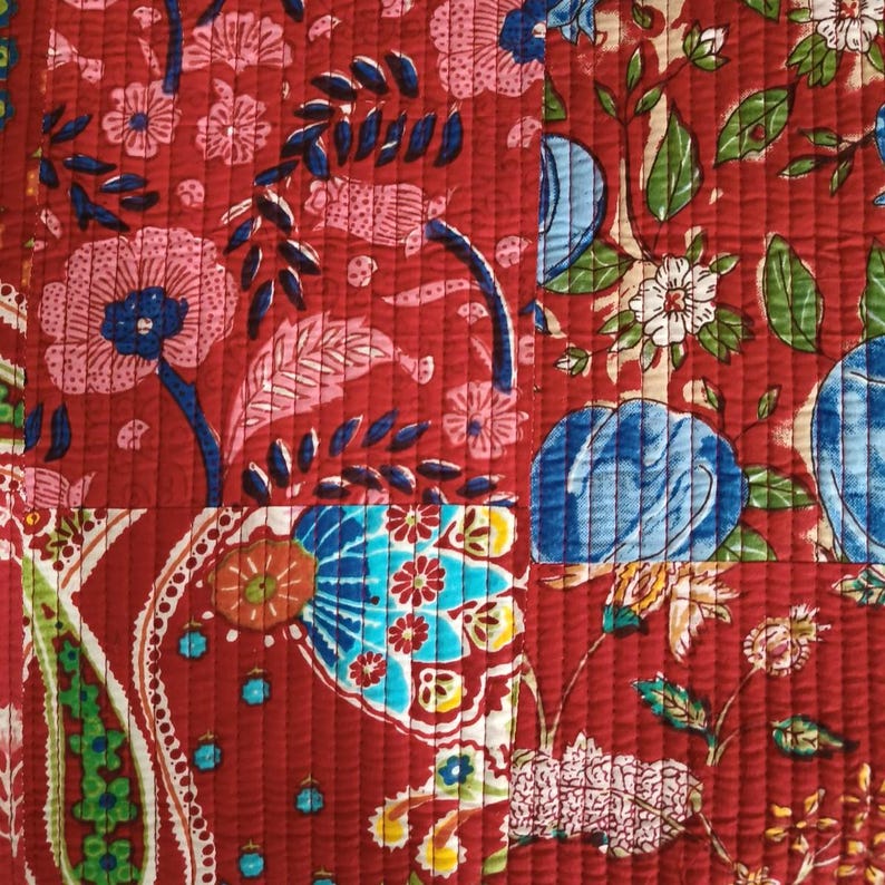 May include: A quilted fabric featuring a vibrant red background with a multi-coloured floral and paisley pattern. The design includes large pink flowers, blue circular motifs, and green leaves, all stitched with horizontal lines across the surface.