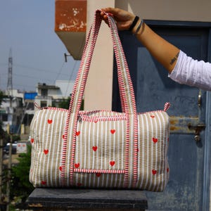 May include: A quilted duffel bag featuring a beige and white vertical striped pattern with small red heart motifs. The bag includes two long shoulder straps with red and white striped trim, a top zipper closure, and a front pocket with a zipper.