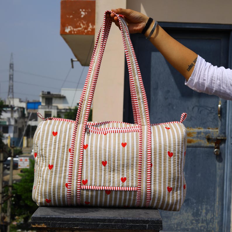 May include: A quilted duffel bag featuring a beige and white vertical striped pattern with small red heart motifs. The bag includes two long shoulder straps with red and white striped trim, a top zipper closure, and a front pocket with a zipper.