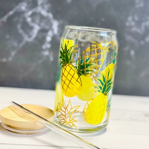 Może przedstawiać: Szklany kubek do picia z pokrywką i słomką. Kubek ma wzór ananasa z żółtymi i zielonymi ananasami na białym tle.