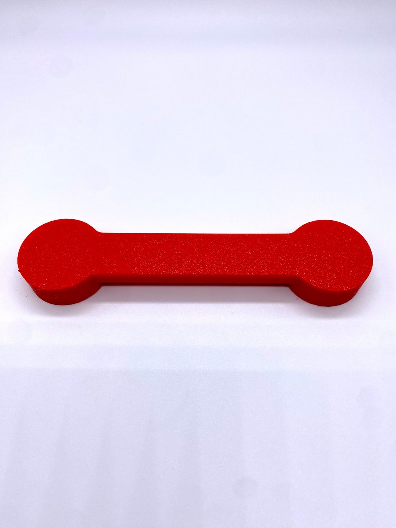 May include: A red, cylindrical phone holder with rounded ends. The holder is made of a smooth, solid material.