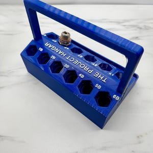 以下が含まれることがあります： 青い3Dプリントの工具ホルダー。ハンドル付き。ホルダーには、数字と文字でラベル付けされた複数の六角形の穴があります。「THE PROJECT HANGAR」の文字が側面に印刷されています。金属製の継手が穴の1つに挿入されています。