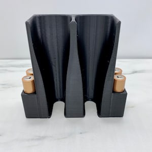 May include: A black 3D printed battery holder with two compartments for storing batteries. The holder has a textured surface and is designed to hold AA batteries. Four batteries are visible in the holder.