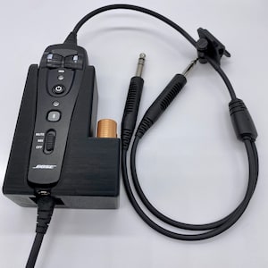 May include: A black Bose microphone with a mute button, a volume knob, and a power button. The microphone is connected to a black cable with a 1/4 inch jack. The microphone is also connected to a black cable with a 3.5 mm jack. The microphone is sitting on a black surface.