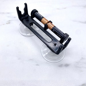 May include: A black 3D printed battery holder with two suction cups. The holder is designed to hold two AA batteries. The batteries are Duracell brand and are visible in the holder.