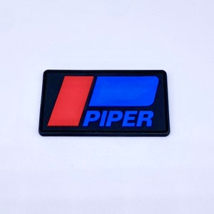 May include: A rectangular black emblem with the word "PIPER" in blue, and a red and blue design. The emblem is on a white surface.