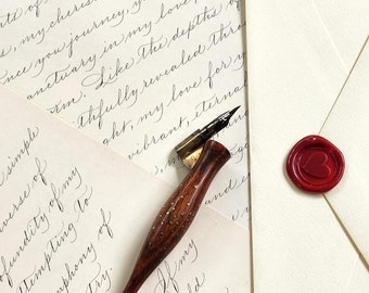 May include: A wooden calligraphy pen with a brass nib and a red wax seal with a heart design on a white envelope. The pen is resting on a piece of paper with handwritten text.