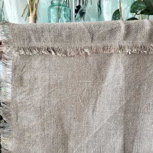 May include: Close-up of a natural-colored linen fabric with a fringed edge. The fabric has a textured appearance and a neutral, earthy tone. The fringe adds a rustic detail to the textile.