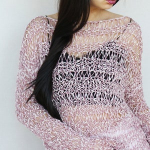 May include: A light pink open-weave sweater with long sleeves. The sweater has a loose, airy design, revealing a black top underneath. The knitwear is a blend of pink and white threads, creating a textured look. The garment is part of a fashion ensemble.