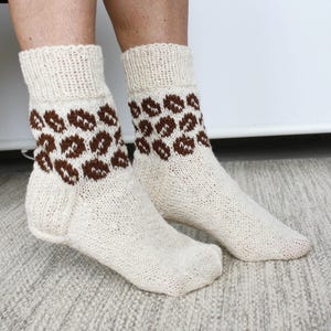 May include: A pair of cream-colored knit socks featuring a brown leopard print pattern around the ankle area. The socks have a ribbed cuff at the top and a plain knit foot section, crafted from a textured yarn with visible stitch details.