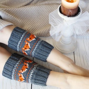以下が含まれることがあります： オレンジ色のキツネ模様と白いアクセントが付いた、グレーのニットレッグウォーマーのペア。 レッグウォーマーは、背景に火の灯ったキャンドルを添えて、明るい木製の表面に表示されています。