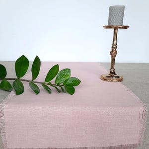 Peut inclure: Chemin de table en lin rose poudré avec bordure effilochée. Un bougeoir doré supporte une bougie grise texturée. Une branche de feuilles vertes repose sur le chemin de table. Le chemin de table mesure environ 30 cm de large.