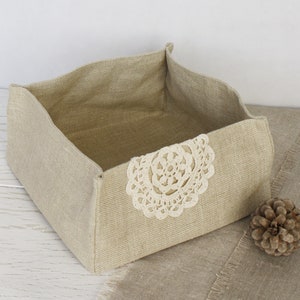 May include: A beige linen storage basket with a cream-colored lace applique on the front. The basket has a rectangular shape with folded-over edges. A pinecone rests on the surface next to the basket.