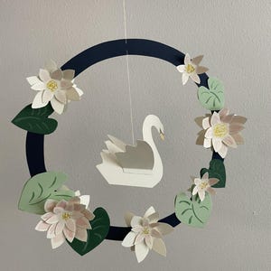 Op de afbeelding: Een handgemaakte mobiel met een witte zwaan die hangt in een marineblauwe ring versierd met papieren bloemen en groene bladeren. De bloemen hebben roze en gele accenten, waardoor een delicate, door de natuur geïnspireerde decoratie voor de kinderkamer ontstaat.