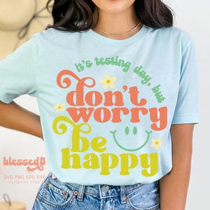 May include: A light blue t-shirt with a retro-style graphic that says "It's testing day, but don't worry be happy". The graphic is in shades of orange, yellow, and green with a smiley face and flowers.