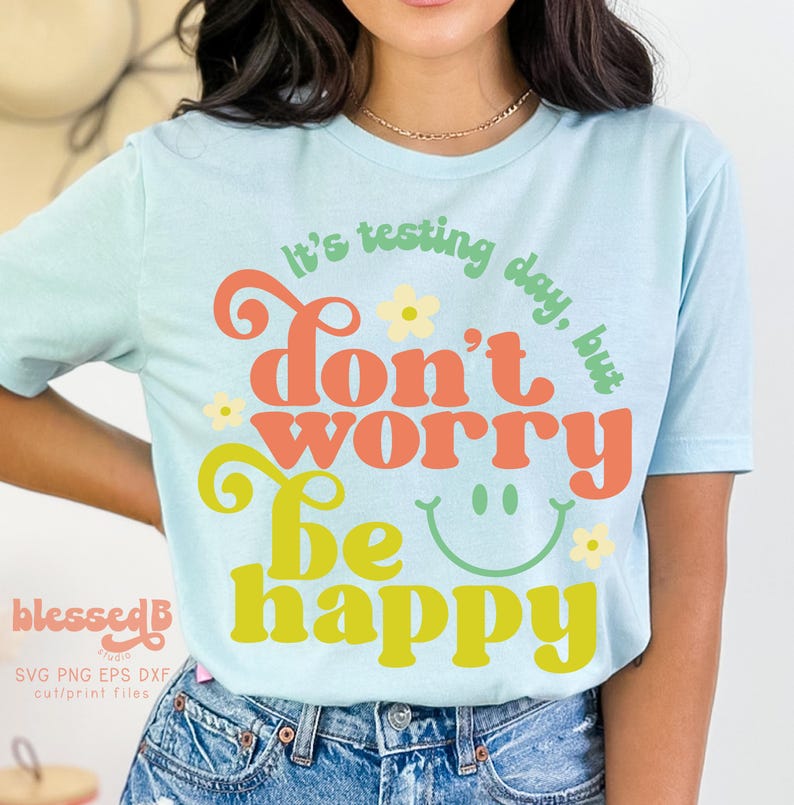 May include: A light blue t-shirt with a retro-style graphic that says "It's testing day, but don't worry be happy". The graphic is in shades of orange, yellow, and green with a smiley face and flowers.