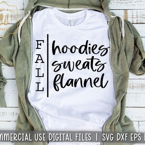 Puede incluir: Camiseta blanca con texto negro que dice "FALL hoodies sweats flannel". La camiseta est&aacute; doblada y colocada sobre una superficie de madera con una sudadera con capucha verde drapeada sobre ella.