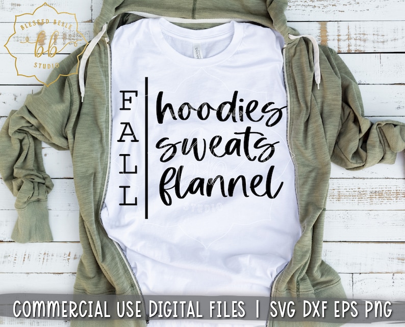 Puede incluir: Camiseta blanca con texto negro que dice "FALL hoodies sweats flannel". La camiseta est&aacute; doblada y colocada sobre una superficie de madera con una sudadera con capucha verde drapeada sobre ella.