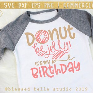 Op de afbeelding: Een wit T-shirt met een grijze mouw, met een roze en bruine donut-afbeelding met de tekst "Donut be jelly! It's my Birthday".
