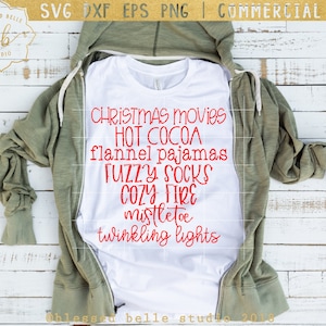 May include: A white t-shirt with red text that reads "Christmas movies, hot cocoa, flannel pajamas, fuzzy socks, cozy fire, mistletoe, twinkling lights". The t-shirt is laying on a wooden surface with a green hoodie on top of it.