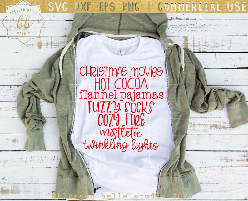 May include: A white t-shirt with red text that reads "Christmas movies, hot cocoa, flannel pajamas, fuzzy socks, cozy fire, mistletoe, twinkling lights". The t-shirt is laying on a wooden surface with a green hoodie on top of it.
