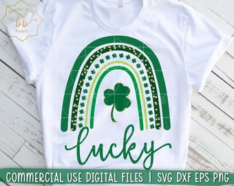 May include: A white t-shirt with a green rainbow design and a green shamrock. The rainbow is decorated with gold glitter and green shamrocks. The text "Lucky" is written in green cursive below the rainbow.
