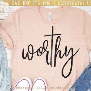 May include: A pink t-shirt with the word "worthy" written in black cursive lettering with a cross above the word.