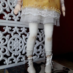 May include: A handmade doll with a yellow dress, white lace trim, white tights, and brown boots. The doll is standing on a book.