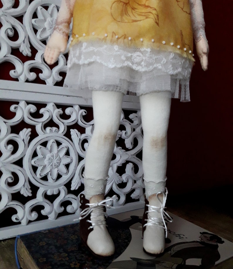 May include: A handmade doll with a yellow dress, white lace trim, white tights, and brown boots. The doll is standing on a book.