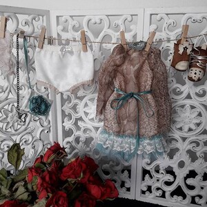 May include: A white lace-trimmed undergarment, a brown and white patterned dress with a teal ribbon, and a pair of brown and white boots hang on a clothesline against a white, ornate background.