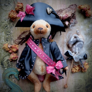 Könnte beinhalten: Ein handgefertigter Teddybär, der eine schwarze Jacke mit rosa Akzenten, eine rosa Schärpe und einen schwarzen Hut trägt. Der Bär hat ein hellbraunes Gesicht und einen hellbraunen Körper. Der Hut ist mit einer Schleife und einem herzförmigen Schmuck verziert. Getrocknete Rosen sind im Hintergrund.