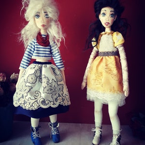 May include: Two handmade dolls with painted faces and detailed clothing. The doll on the left has white hair, a blue and white striped shirt, a white skirt with gold embroidery, and blue shoes. The doll on the right has black hair, a yellow dress with brown trim, and white shoes.