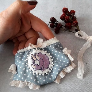 May include: A light blue fabric face mask with a white lace trim and a purple embroidered dinosaur design. The mask has white polka dots and a white ribbon.