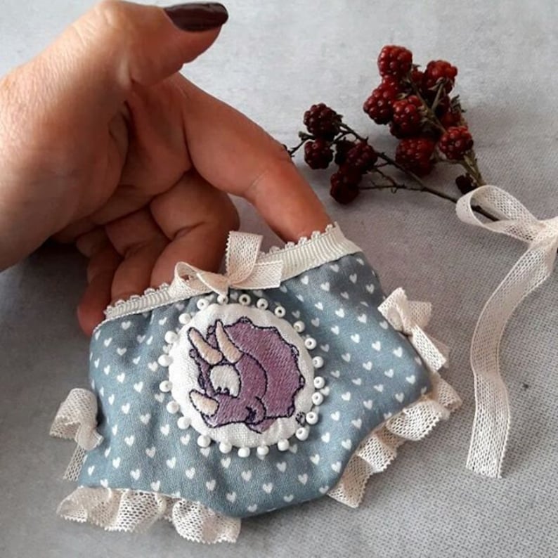 May include: A light blue fabric face mask with a white lace trim and a purple embroidered dinosaur design. The mask has white polka dots and a white ribbon.