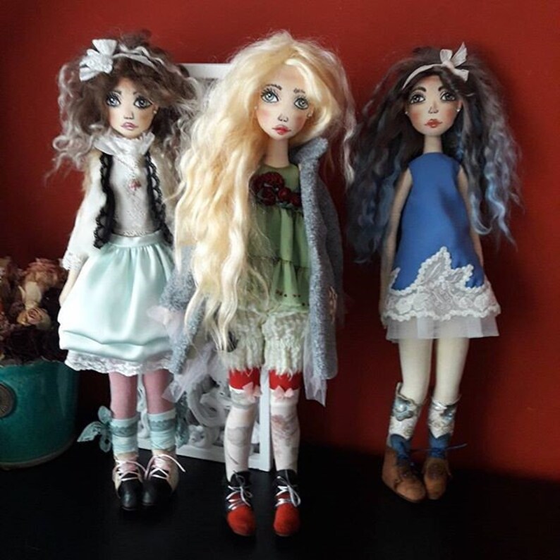 May include: Three handmade fabric dolls with long, curly hair. The dolls are wearing different outfits, including a blue dress, a green dress with a gray cardigan, and a white dress with a black vest. The dolls are standing on a black surface.