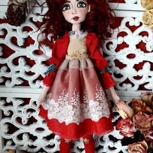 May include: A handmade doll with red curly hair, wearing a red dress with a white lace trim and brown boots. The doll is standing in front of a white decorative screen.