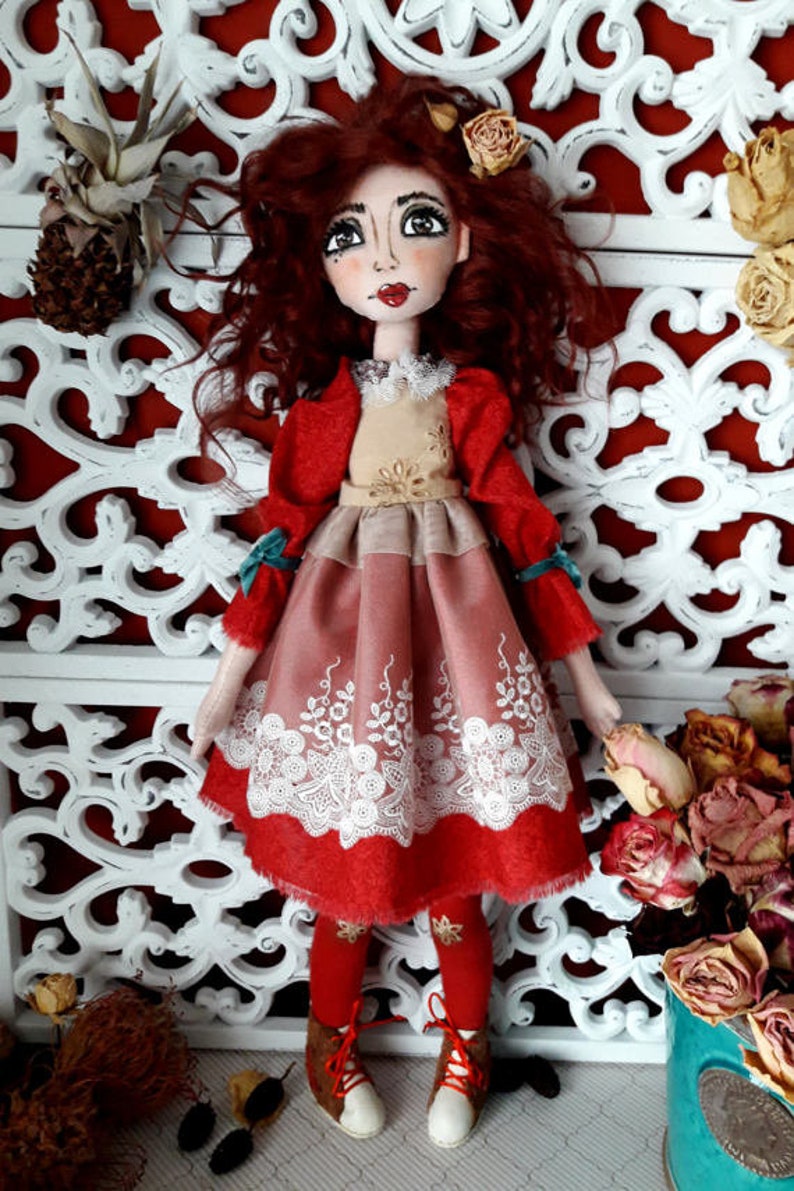 May include: A handmade doll with red curly hair, wearing a red dress with a white lace trim and brown boots. The doll is standing in front of a white decorative screen.